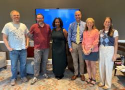 Group photo of six faculty members standing indoors in front of a screen. They are smiling and dressed casually to business casual, representing the Faculty Grant Cohort 2025. The room has a patterned carpet and neutral walls.
