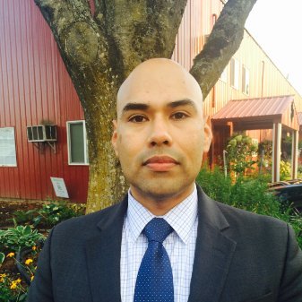 - Man in a suit and tie standing outdoors in front of a red building with a tree and greenery in the background.