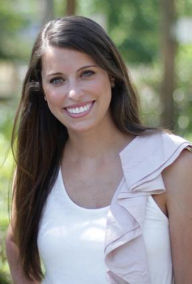 - A woman with long brown hair smiles while standing outdoors, wearing a white sleeveless top with a ruffled detail on one shoulder.