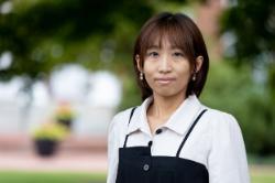 Shimeng Wang - Woman with short brown hair wearing a white collared shirt and black dress, standing outdoors with greenery and blurred flowers in the background.