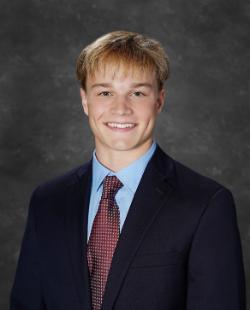 A young man in a dark suit jacket, light blue shirt, and patterned maroon tie smiles in a formal studio portrait with a gray textured background.
