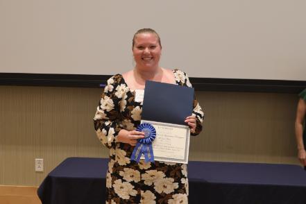 - A Longwood graduate smiles while holding a certificate and first-place award ribbon.