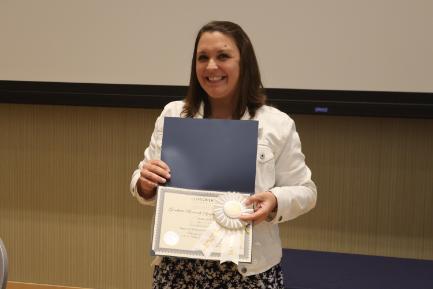 A Longwood graduate smiles while holding a certificate and award ribbon.