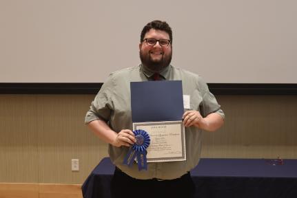 - A Longwood graduate smiles while holding a certificate and first-place award ribbon.