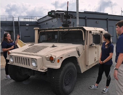 Joe Gills and Tank - Three people stand beside a beige military vehicle equipped with mounted technology. The group wears navy shirts and appears to be discussing the equipment. The scene takes place on a paved lot outside a gray industrial building. Cloudy skies and soft lighting create a neutral, professional setting.