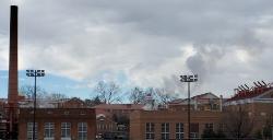 - A cluster of brick campus buildings sits beneath a cloudy sky. Steam or smoke rises behind the rooftops near tall stadium lights. A red smokestack stands on the left side of the scene. Bare winter trees frame the view across the campus landscape.