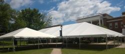 - Two large white event tents stand on a grassy lawn under a bright sky. A brick campus building with columns rises in the background. A lamppost sits centered between the tent structures. Trees and shadows frame the open, outdoor gathering space.