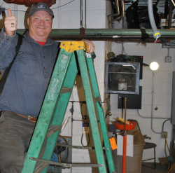 - A worker stands on a green ladder inside an industrial mechanical room. He smiles and gives a thumbs-up while holding onto the ladder. Pipes, ducts, and equipment fill the dim workspace around him. Tools, cables, and a trash bin sit on the floor behind the ladder.