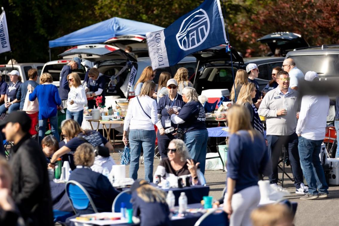 Homecoming Lancer Family Tailgate - Alumni gather for a tailgate during Longwood University Homecoming weekend. Groups stand and sit around tables with food and drinks in a parking lot. A blue Longwood flag waves above tents and open car trunks. People wear Longwood apparel and socialize in a festive outdoor setting.
