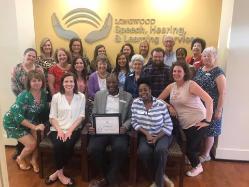- Large group of faculty, staff, and students pose together inside Longwood University’s Speech, Hearing, and Learning Services clinic. A man seated in the center holds a framed certificate while others stand around him smiling. The program’s logo and “Speech, Hearing, & Learning Services” sign are displayed on the wall behind the group. Photo appears to mark a recognition or celebration within t