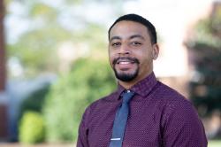 Quincy Goodine - A man smiles in a professional headshot outdoors. He wears a maroon patterned button-down shirt with a blue tie and tie clip. He faces the camera with a neatly trimmed beard and short hair. Trees and campus buildings blur softly in the background.