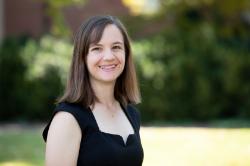 Dr. Sarai Blincoe - Portrait of a woman smiling outdoors in natural light. She has shoulder-length brown hair and wears a black sleeveless top. A small necklace rests at her collarbone. Green trees and lawn blur softly in the background.