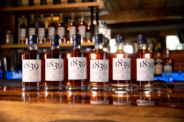 Bottles of 1839 Bourbon - Six bottles of 1839 Collection single barrel bourbon whiskey sit on a wooden bar. The amber liquid glows under warm lighting inside a distillery. Shelves of additional bottles line the blurred background. Each label reads “1839 Collection” and “2023 Reserve.”
