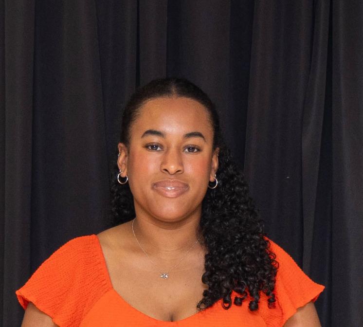 A woman in an orange top poses for a headshot in front of a black curtain.