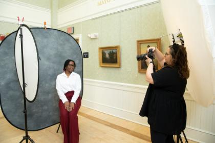 Career Cafe 3 - A student poses for a professional headshot against a gray backdrop while a photographer takes the picture using studio lighting and camera equipment in a room decorated with framed artwork.