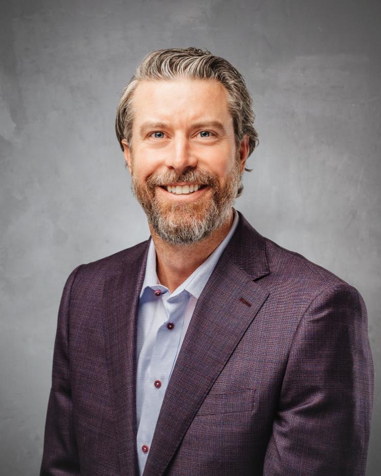 Chris Isaacs headshot - A man with short gray hair and a trimmed beard smiles while wearing a light blue button-down shirt and a maroon plaid blazer against a gray studio background.