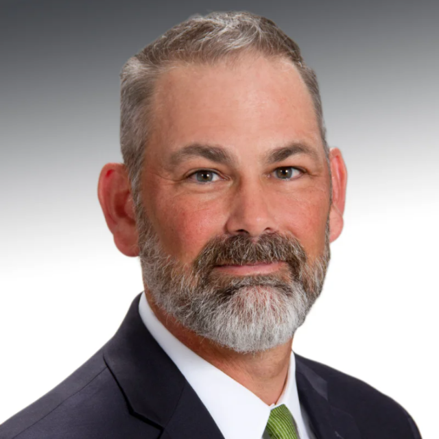 Tage Counts headshot - Professional headshot of a man with short gray hair and a trimmed salt-and-pepper beard, wearing a dark suit, white shirt, and green tie against a gradient gray background.