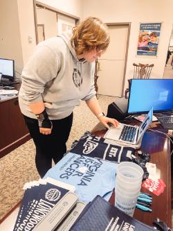Spin for Swag - A person stands at a desk working on a laptop in an office setting.   They wear a light hoodie and a wrist brace, surrounded by promo items.   T-shirts, cups, and folders display “VIRGINIA LANCERS” and “LONGWOOD UNIVERSITY.”   The background includes monitors, furniture, and a poster about career advancement.