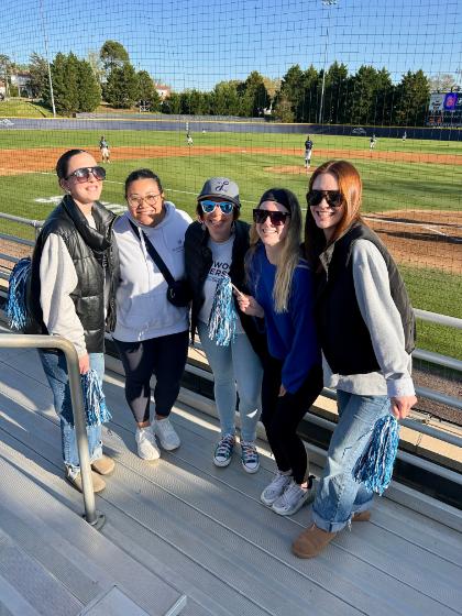 Baseball Night - Five people stand on bleachers at a baseball field holding pom-poms. They wear casual clothes and sunglasses, showing team spirit. The field, players, scoreboard, and trees are visible in the background. The scene captures enthusiastic spectators enjoying the game.