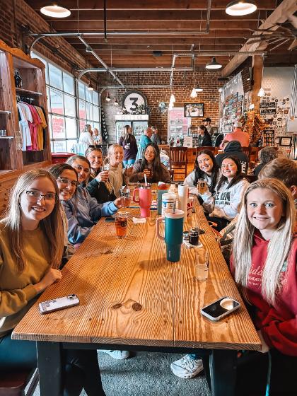 Brewery Night - A group of people sit around a long wooden table in a rustic café.   The space features brick walls, wooden beams, and decorative shelves.   They appear to be enjoying drinks and smiling in a relaxed setting.   Natural light enters through large windows on the left side.