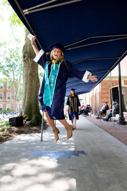 graduate jumps after receiving her diploma.  - A graduate in cap and gown jumps while holding a pole under a canopy.   They wear a teal stole with “ZTA” lettering, celebrating the occasion.   Another graduate walks in the background near brick buildings and trees.   The scene captures a joyful moment in an outdoor graduation setting.