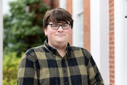 Person wearing glasses and a black-and-olive checkered button-down shirt stands outside a brick building. They are centered in the frame, looking at the camera with a neutral expression. Green foliage and blurred brick architecture create a soft background. Natural lighting highlights the subject’s face and the texture of their shirt.