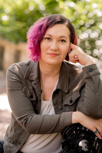 - A person with short, wavy hair dyed pink on one side sits outdoors on a bench. They rest their head lightly on one hand and look toward the camera. They wear an olive-green jacket over a light-colored top. The background is softly blurred greenery, creating a natural portrait setting.