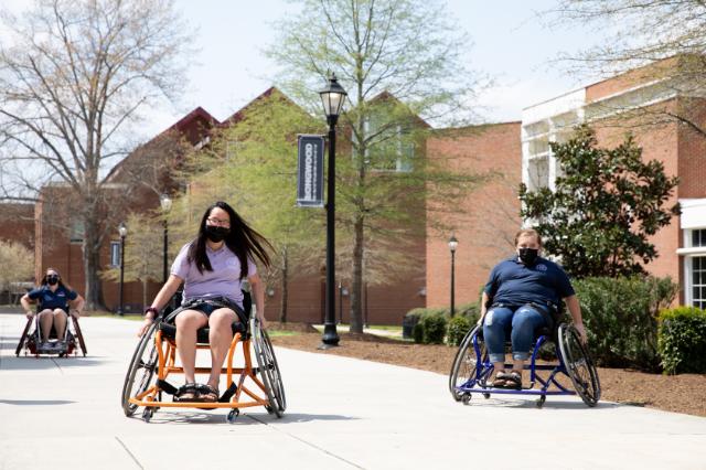 Two students trial sports wheelchairs. - Three students use sport wheelchairs while moving along a campus walkway. They wear face masks and casual university apparel. Brick academic buildings, trees, and lamp posts line the path. A Longwood banner is visible, indicating a university campus setting.