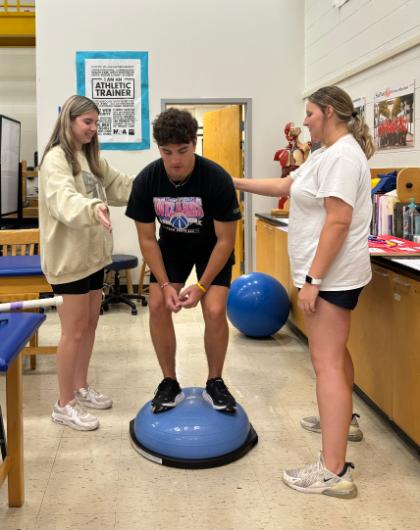 Students working with a Bosu ball in KINS 298 - Three students practice a balance exercise in an athletic training lab. One student stands on a BOSU ball while bending at the knees. Two classmates stand nearby, offering guidance and support. Anatomy models and training equipment are visible in the background.