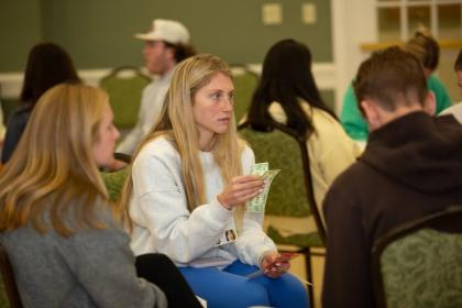 Student participates in the 2025 Poverty Simulation event - A student sits in a circle holding paper money and cards during an activity. She looks focused while listening to others seated nearby. Other participants sit around her in a classroom or meeting room setting. The scene appears to be part of a poverty simulation or role-playing exercise.