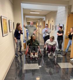 TR students trialing adaptive sports equipment - A group of students gathers in a campus hallway during an adaptive sports activity. Two students use sports wheelchairs at the center of the group. Others stand nearby watching, talking, and offering guidance. Framed artwork lines the hallway walls in an indoor academic building.