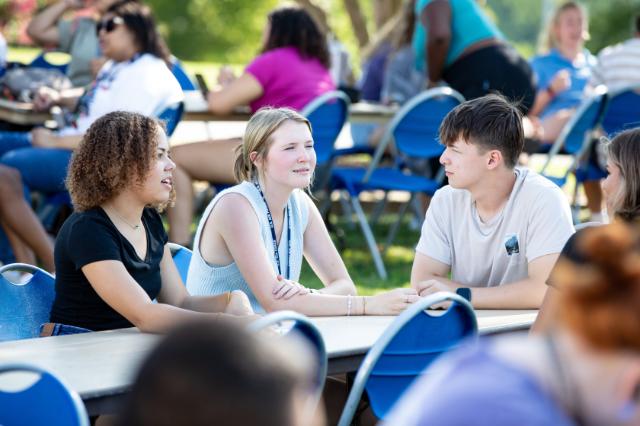 Three students sit at a table and talk. - Three students sit at an outdoor table talking during a campus event. They face each other in conversation, leaning in with relaxed, engaged expressions. Blue folding chairs and tables are arranged across a grassy area. Other students gather and socialize in the background under sunny conditions.