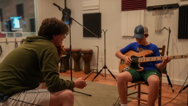 - Instructor sits and listens while a student plays an electric bass guitar.