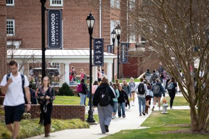 students change classes - Students walk along a busy campus sidewalk between classes. Many carry backpacks as they move through the area. Longwood University banners hang from lampposts along the path. Red-brick buildings and campus landscaping surround the walkway.