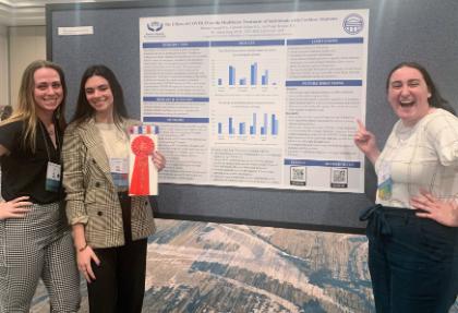 - Three students stand beside a research poster at an academic conference. One student holds a red ribbon indicating a second-place award. The poster discusses the effects of COVID-19 on healthcare treatment for individuals with cochlear implants. The group smiles and points to the poster while celebrating their research recognition.