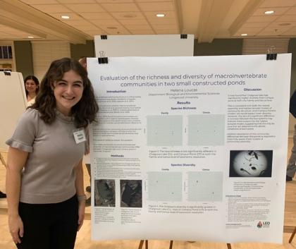 - A student stands beside a research poster at an undergraduate research presentation. The poster is titled “Evaluation of the richness and diversity of macroinvertebrate communities in two small constructed ponds.” Charts and images display results about species richness and diversity. She smiles while presenting her environmental science research.