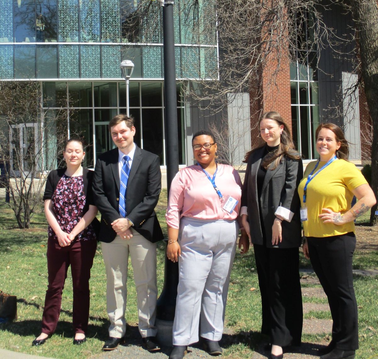 - Five students stand together outdoors in business attire on campus. They pose in front of a modern academic building with large glass windows. Several wear conference or event badges on lanyards. The group smiles for a photo on a sunny day near a campus walkway.