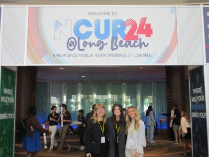 - Three students stand together beneath a sign reading “Welcome to NCUR24 @ Long Beach.” They wear conference badges and smile inside a convention center entrance. Other conference attendees move through the space behind them. Students attend and represent their university at a national research conference.