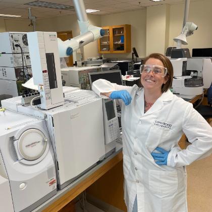 - A researcher wearing safety goggles and gloves stands beside a gas chromatography instrument in a laboratory. She leans on the equipment while wearing a white lab coat labeled Longwood University. Analytical instruments and ventilation arms are visible around the lab workspace. The scene highlights hands-on scientific research using advanced laboratory equipment.