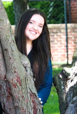 - A student smiles while standing between the branches of a large tree. She leans slightly forward from behind the trunk in an outdoor campus setting. A brick wall and greenery appear softly in the background. Sunlight highlights the tree bark and the student’s long dark hair.