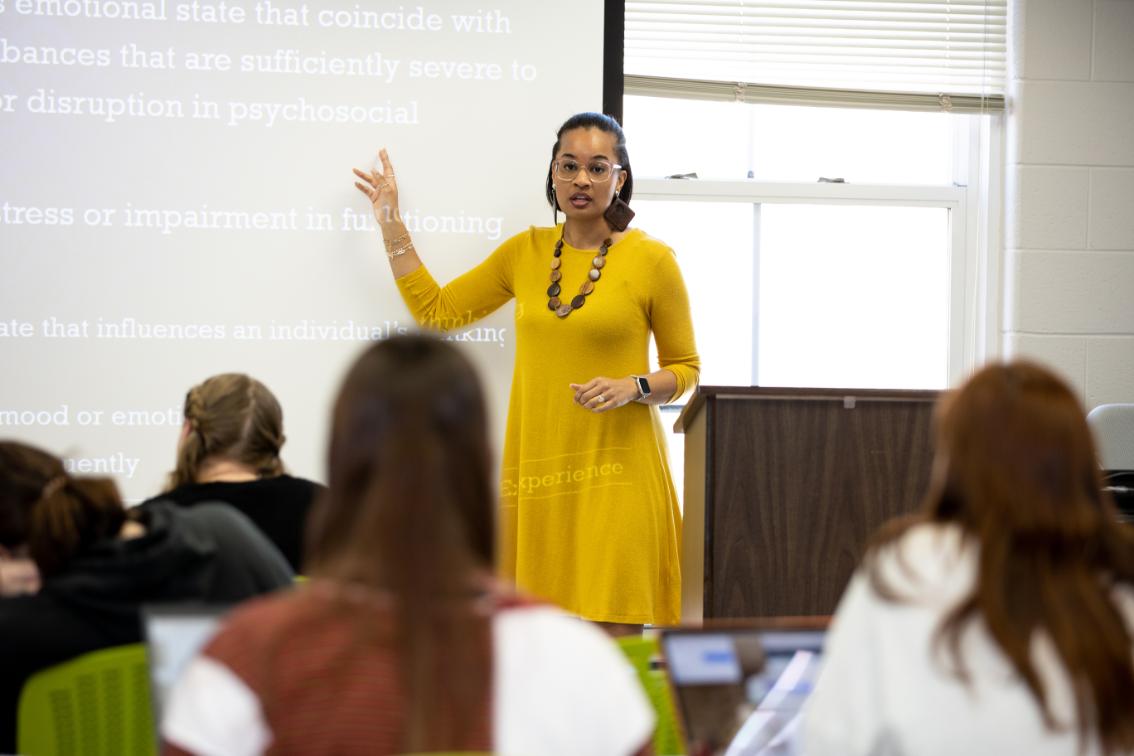 Shayla Walker teaching a class - Professor in a yellow dress lectures at the front of a classroom. She gestures toward projected text displayed on a screen behind her. Students sit facing forward, listening and taking notes. Classroom setting includes a podium, window, and projection screen.
